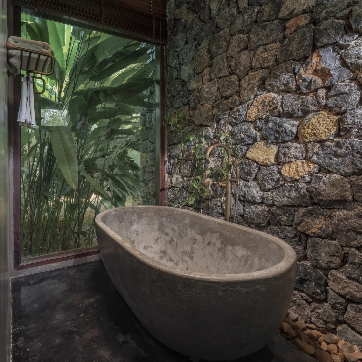 A stone bath stands beside a window filled with tropical foliage