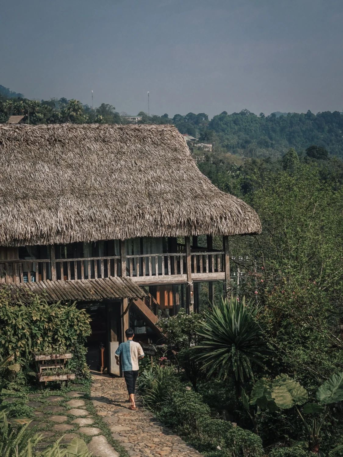 A person walks along a garden path towards a thatched lodge